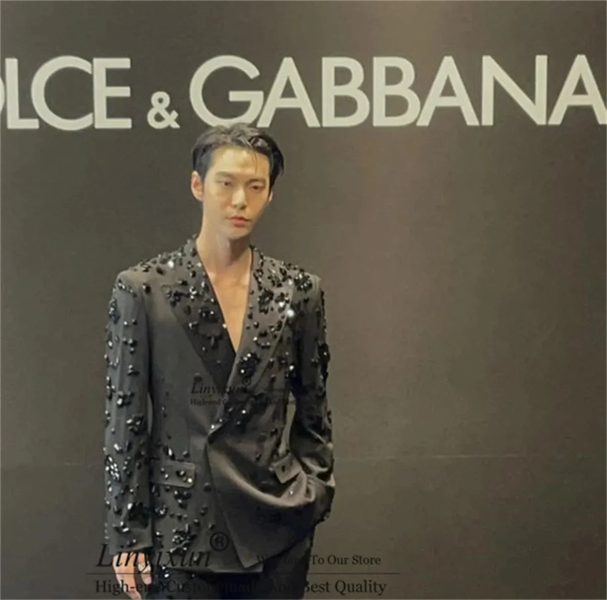 A man wearing a dark gray Dolce & Gabbana blazer embellished with silver studs, standing in front of a dark gray wall with the white Dolce & Gabbana logo.